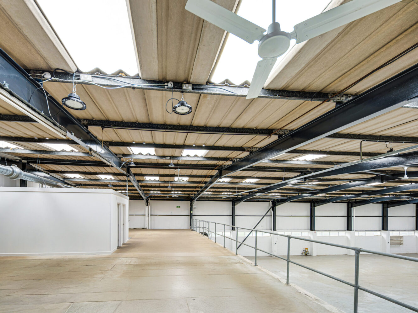 Spacious industrial warehouse interior with concrete floor, metal beams, skylights, ceiling fans, and white partitioned rooms below.
