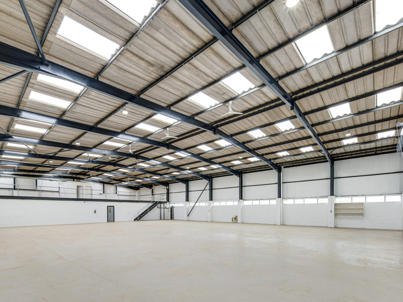 Spacious empty warehouse with a high ceiling, exposed steel beams, skylights, white walls, and a mezzanine with stairs on one side.