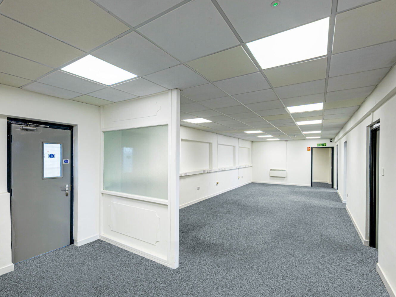 Empty office space with gray carpet, white walls, fluorescent ceiling lights, several doors, and a large interior window partition.