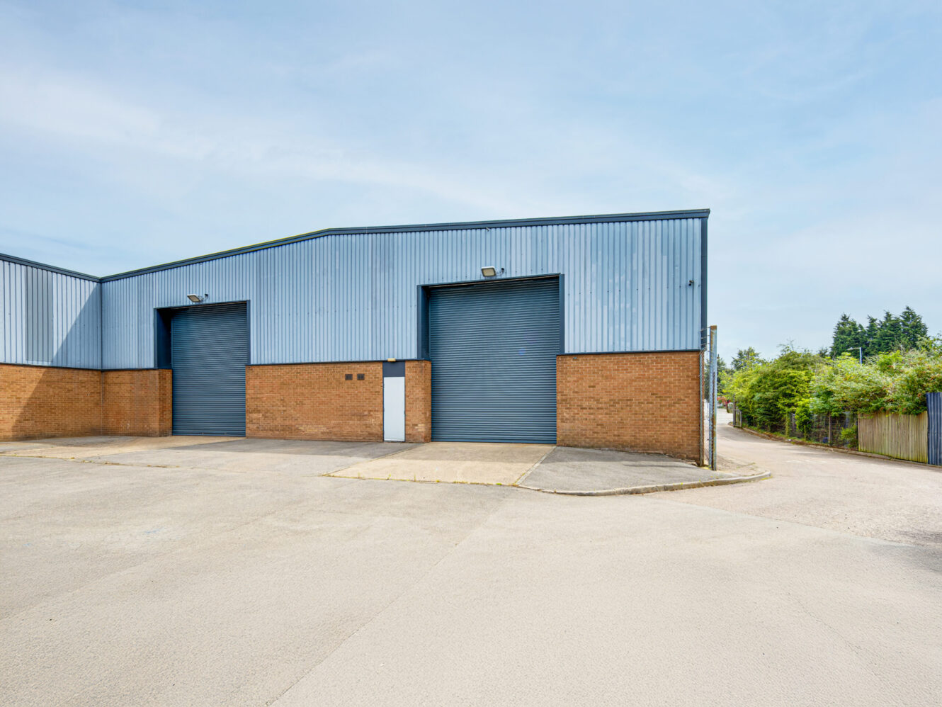 Exterior of a modern industrial warehouse with two large closed roller doors, a small white entry door, and an empty paved area in front.