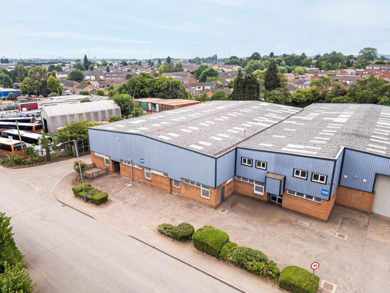 A large industrial warehouse with a blue and brown exterior is situated on a quiet street, with houses and trees visible in the background.
