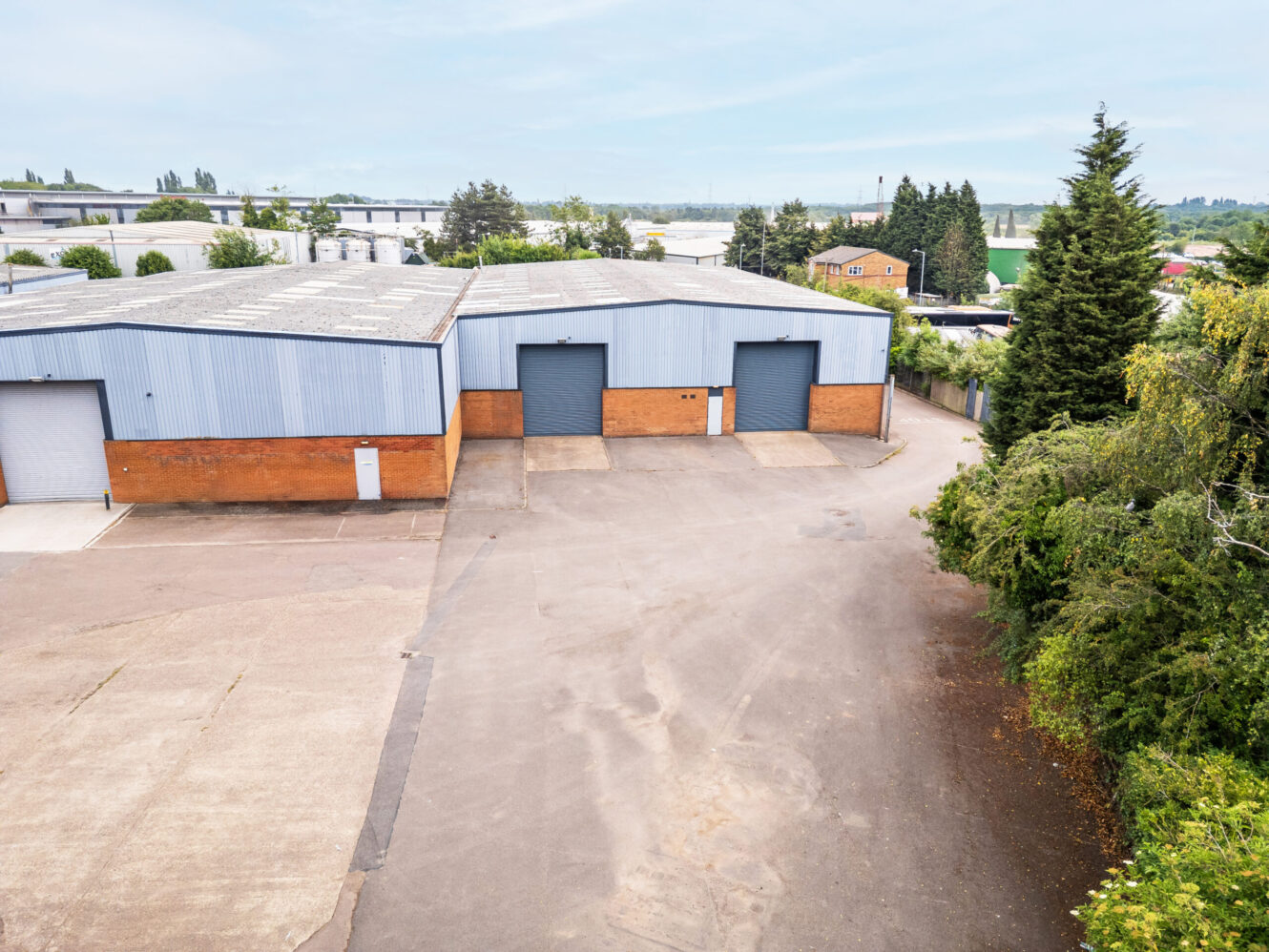 Industrial warehouse with two large roller doors, surrounded by paved area and trees, under a cloudy sky.