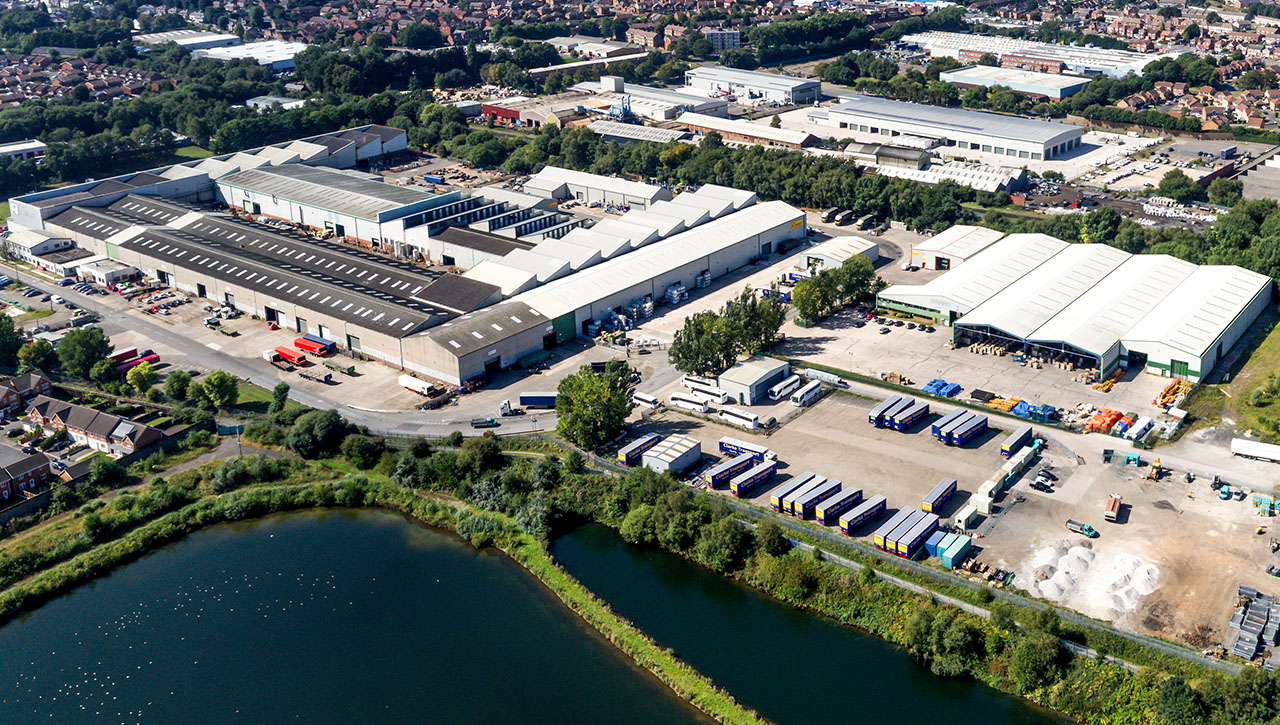 Aerial view of a large industrial complex with multiple warehouses, trucks, and storage areas, situated near a body of water and surrounded by greenery and residential areas.