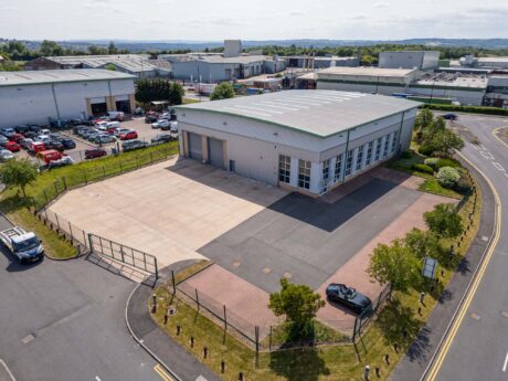 Aerial view of a modern industrial warehouse with parking areas, surrounded by fencing, situated in a business park with other commercial buildings nearby.