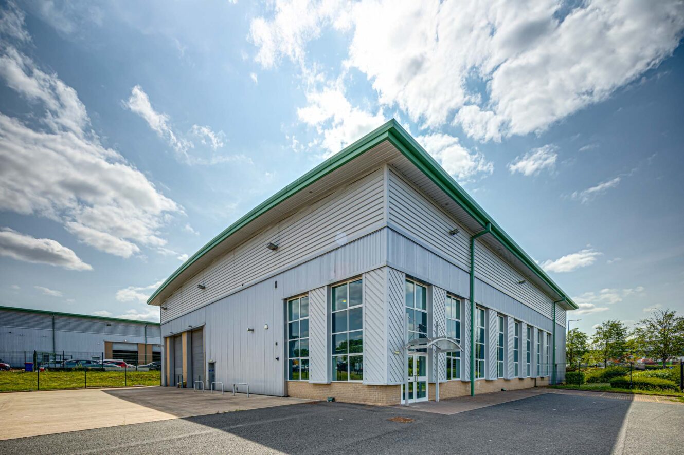 A modern industrial warehouse with large glass windows, metal siding, and a green roof, set against a partly cloudy sky.