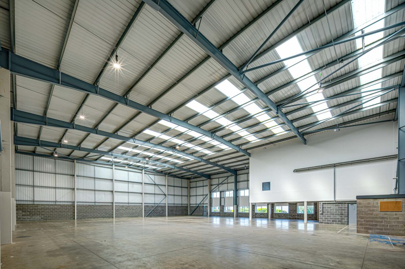 Interior of an empty industrial warehouse with concrete floors, high ceilings, exposed beams, and skylights allowing natural light inside.