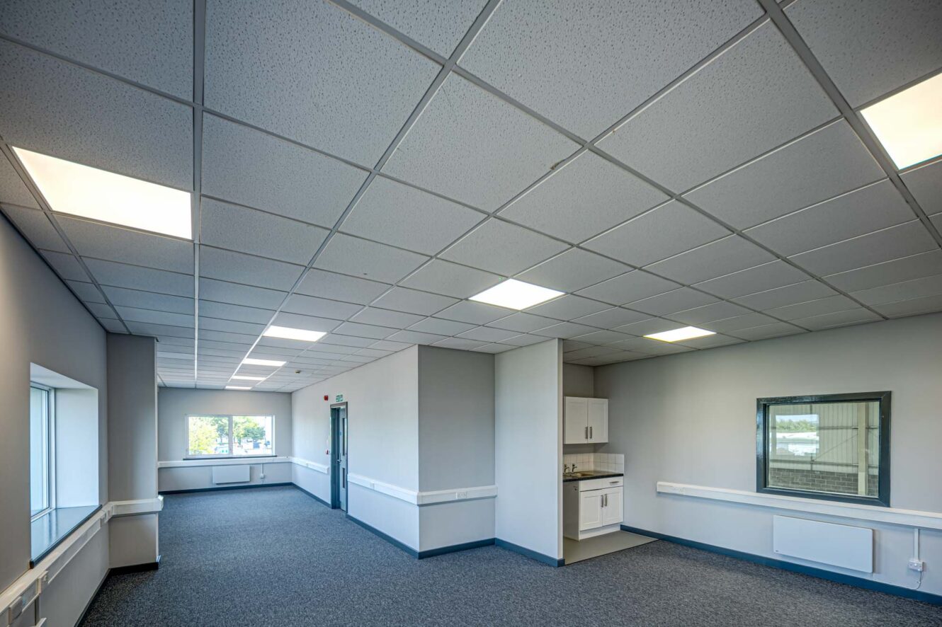 An empty office room with gray carpet, white walls, a drop ceiling with fluorescent lights, large windows, and a small kitchenette area with a sink and cabinets.