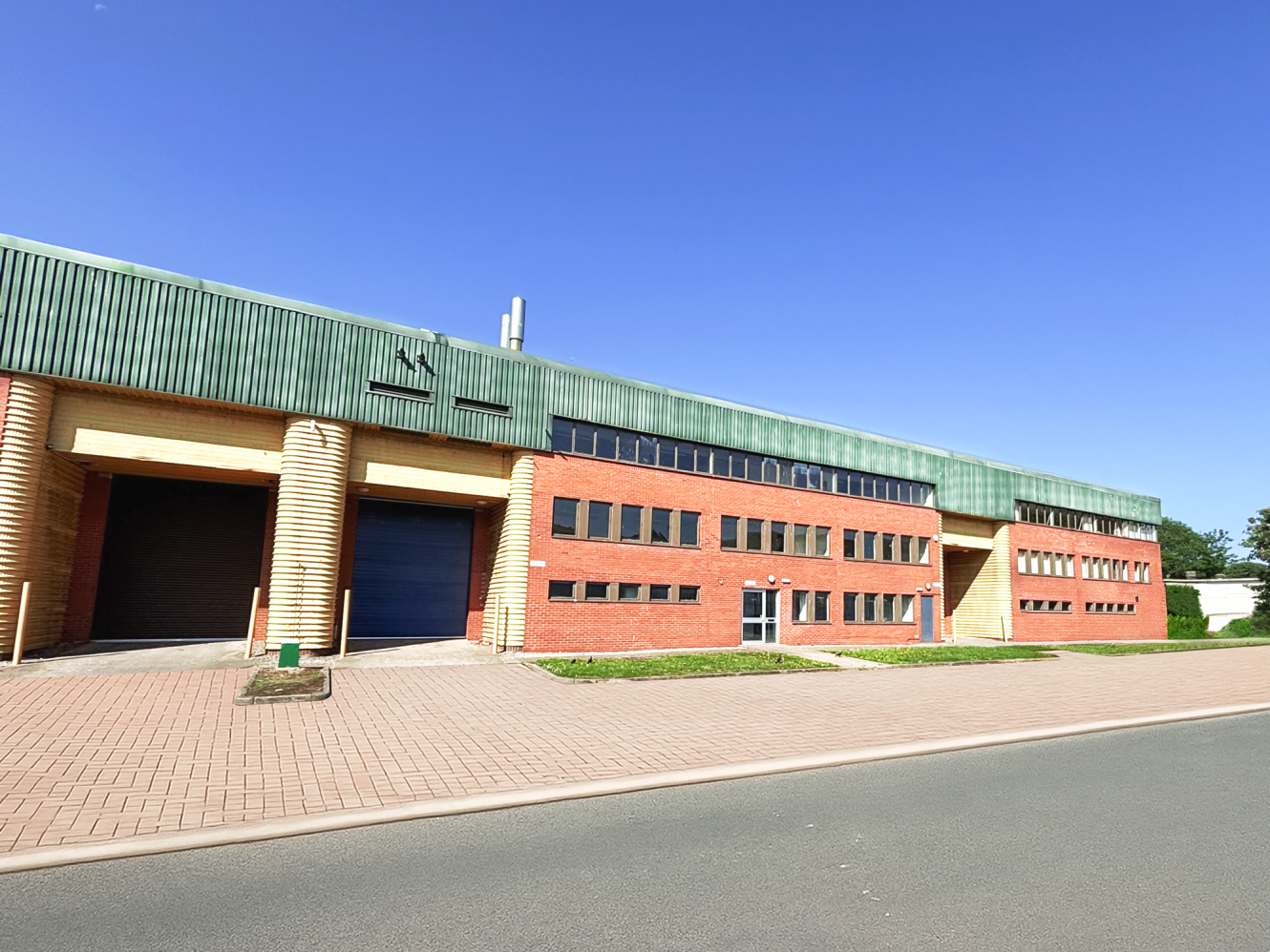 A large industrial building with a green upper section and red brick lower section, featuring multiple windows and a roller shutter door, under a clear blue sky.