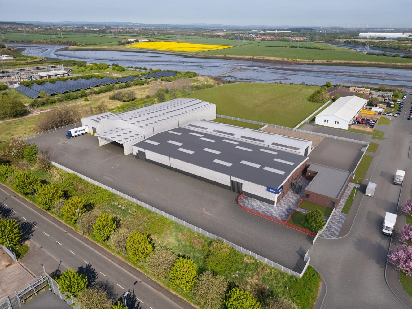 Aerial view of an industrial facility with large warehouse buildings, parking areas, greenery, and nearby river, fields, and solar panels.