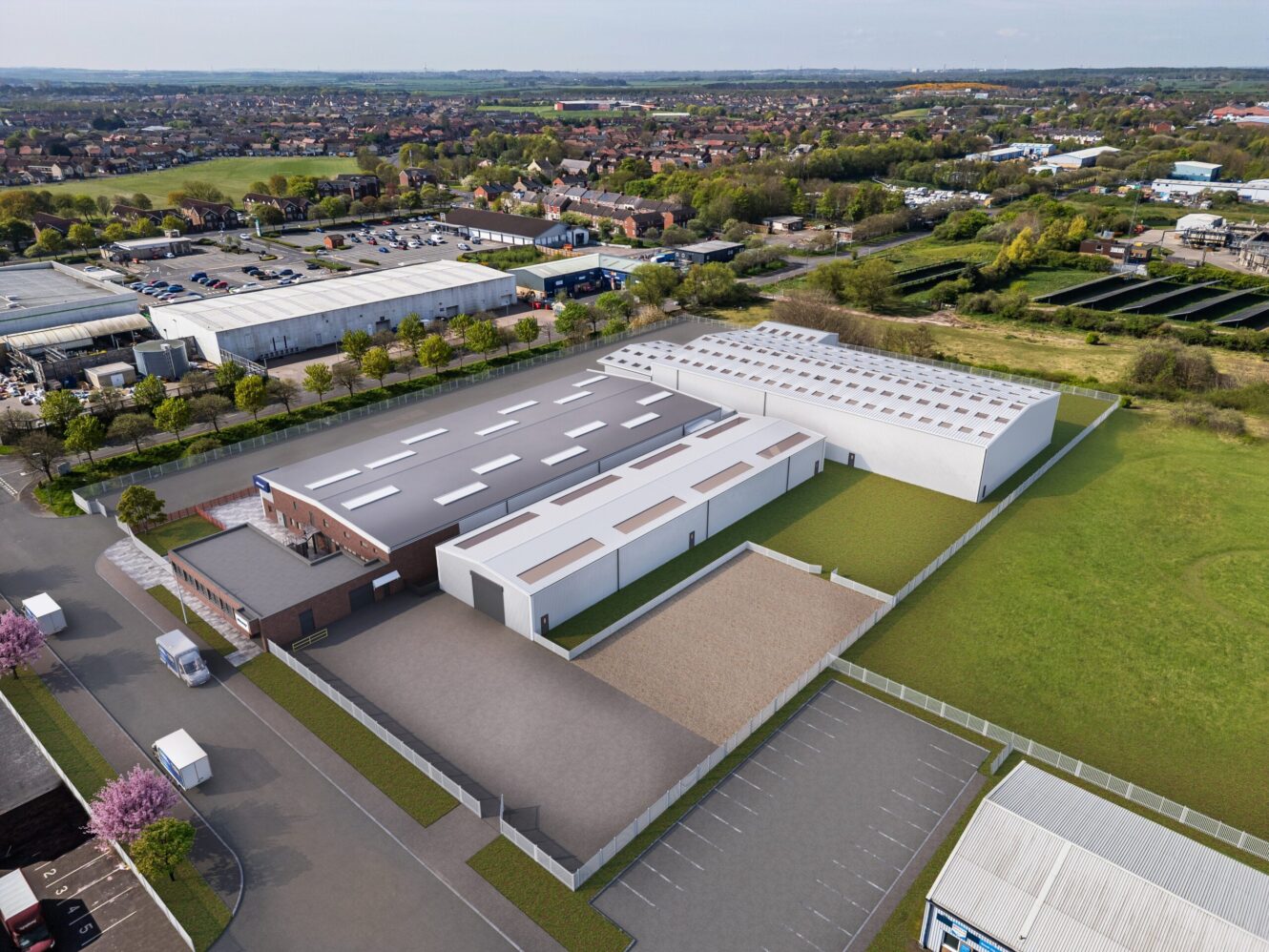 Aerial view of a large industrial building with adjacent car park and fenced area, surrounded by fields and nearby commercial buildings.