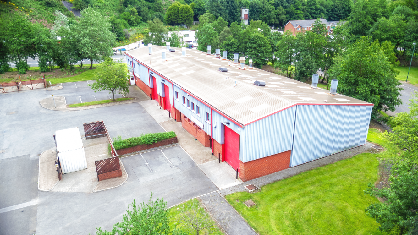 Aerial view of a large industrial warehouse with red and grey walls, surrounded by trees, parking spaces, and some fencing.