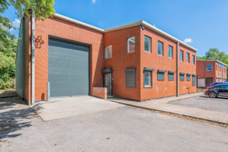 A modern two-storey brick commercial building with large windows, a grey roller shutter garage door, and a small car park in front under a clear blue sky.