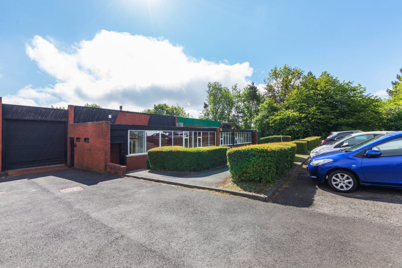 Single-storey brick office building with large windows, surrounded by trimmed hedges, a drive, and parked cars under a sunny sky.