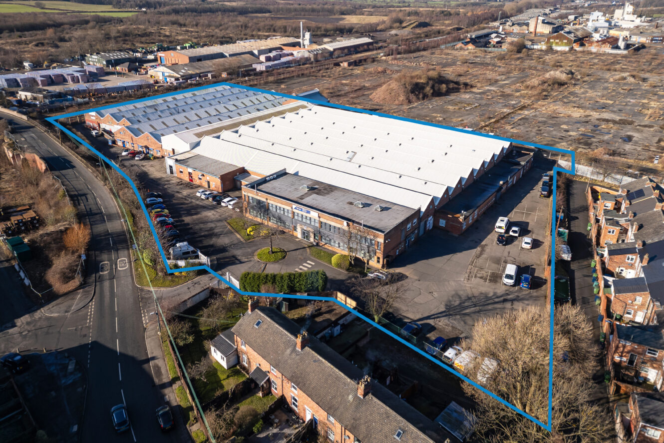 Aerial view of an industrial building with a white roof, car park, and perimeter outlined in blue, surrounded by roads, houses, and vacant land.