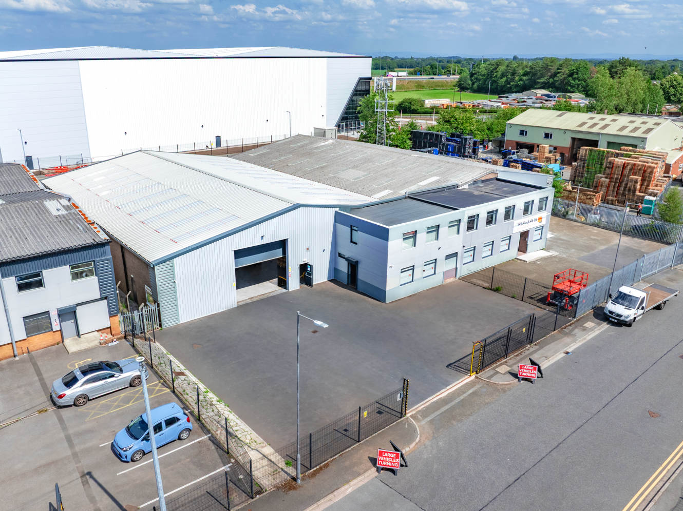 Aerial view of an industrial warehouse building with a fenced car park, several parked vehicles, and surrounding warehouses under a partly cloudy sky.