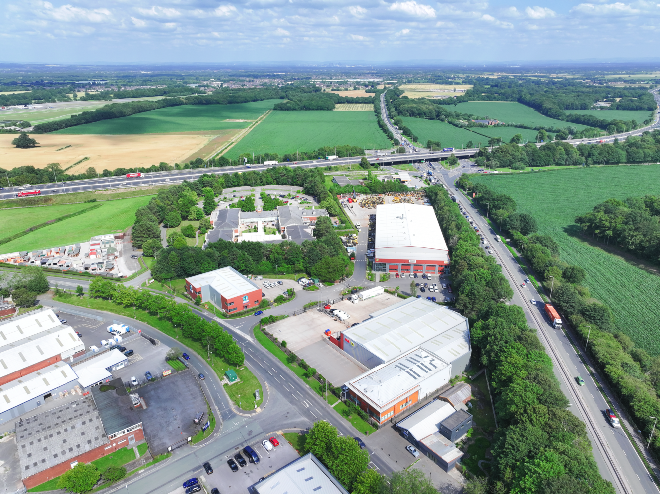 Aerial view of an industrial and office park surrounded by green fields and trees, with several roads and vehicles visible under a partly cloudy sky.