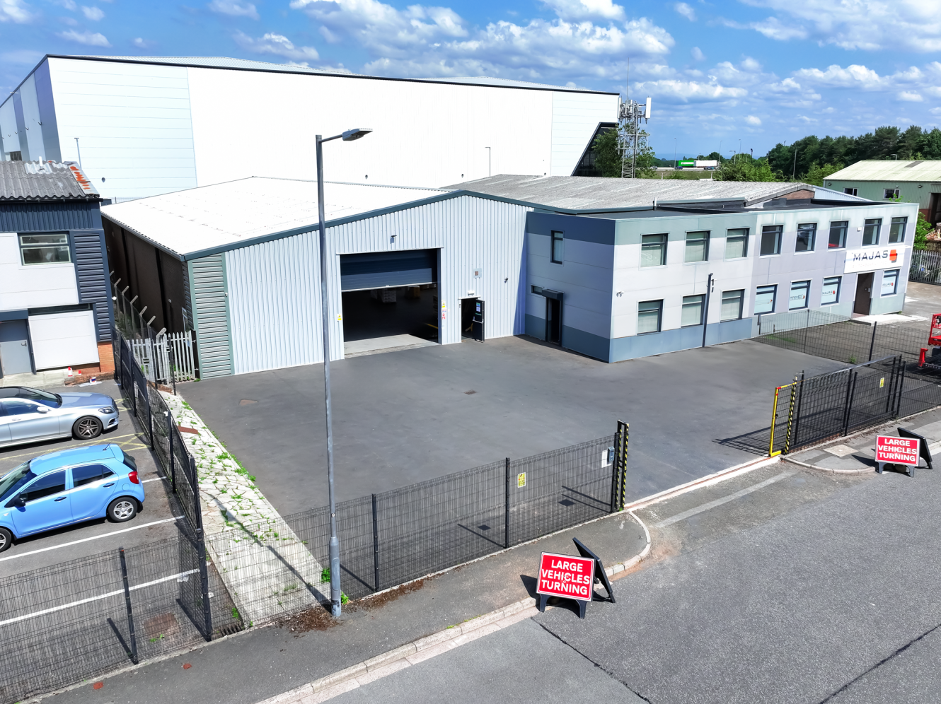 A large industrial building with an attached office section and a fenced car park; signs indicate Large Vehicles Turning near the entrance.