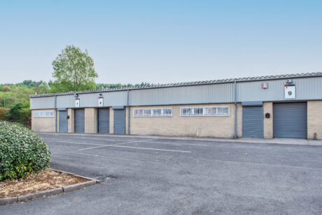 A row of industrial warehouse units with closed roller shutters and numbered signs, adjacent to an empty tarmac car park and a bush in the foreground.