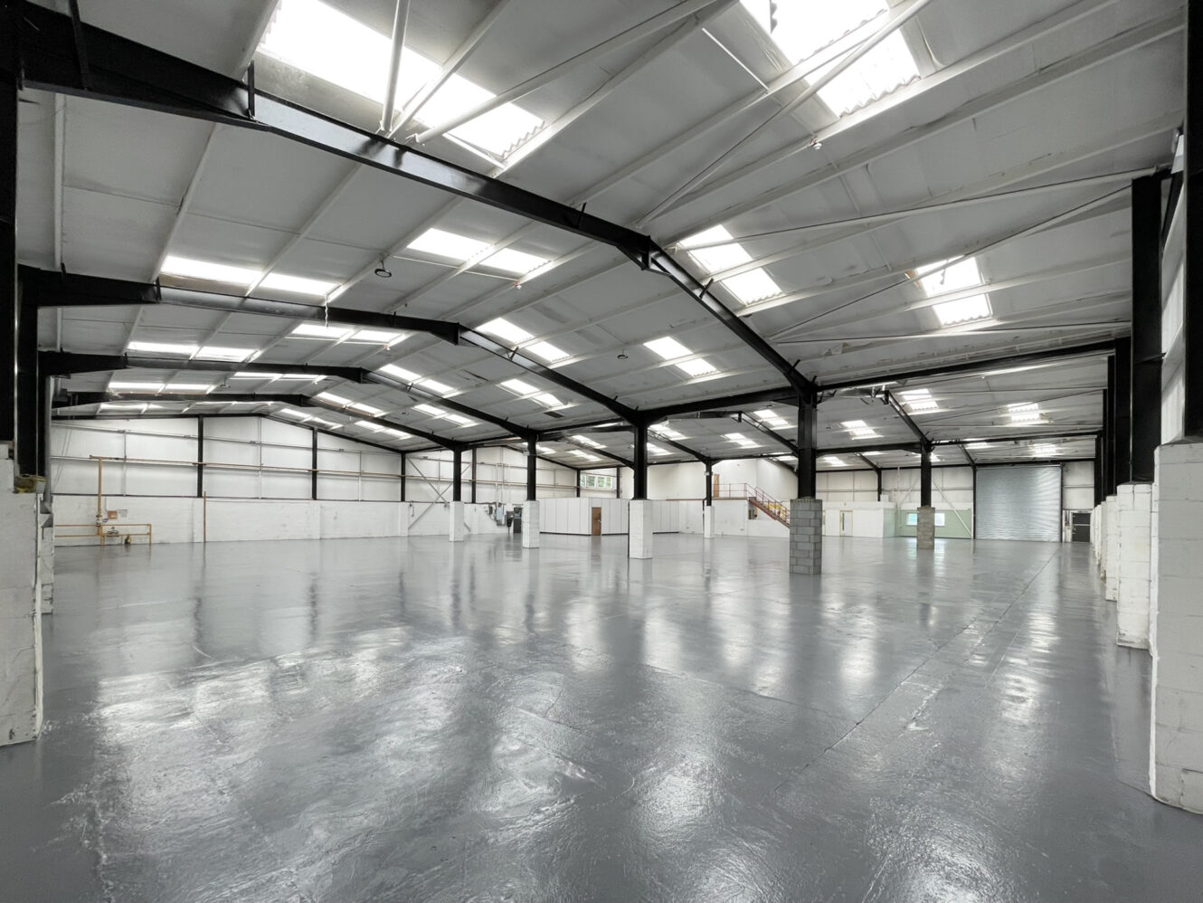 Large, empty warehouse interior with polished concrete floor, white walls, exposed steel beams, and overhead skylights. Some shelving and a staircase are visible along the walls.