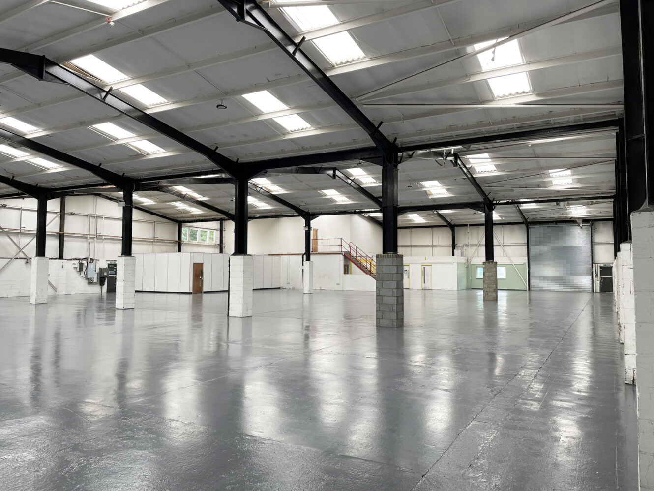 Interior of an empty industrial warehouse with high ceilings, exposed beams, white walls, and polished concrete floors. A staircase leads to a mezzanine level in the background.