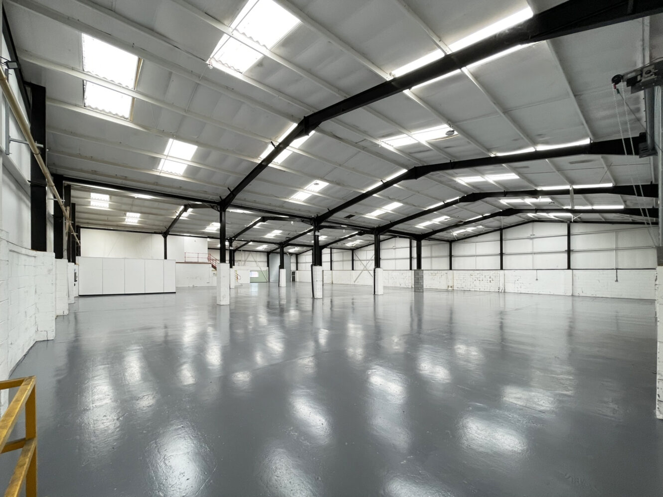 A large, empty warehouse space with white walls, a glossy grey floor, and a high ceiling supported by black steel beams, lit by overhead fluorescent lights.