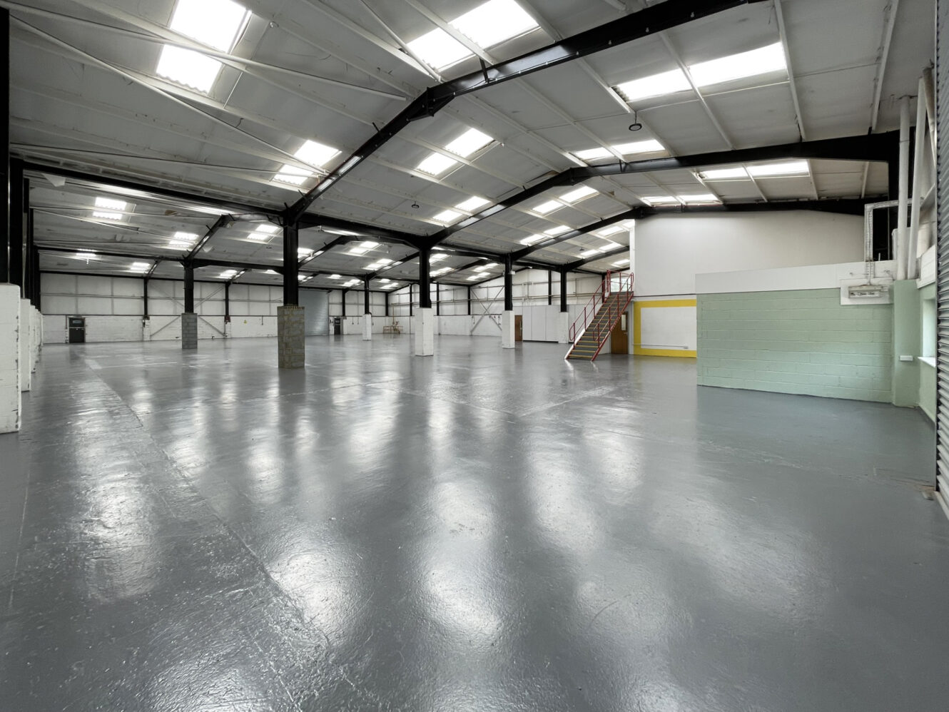 Large, empty industrial warehouse with polished grey floor, white walls, high ceiling, and skylights; partial interior rooms and a red staircase are visible.