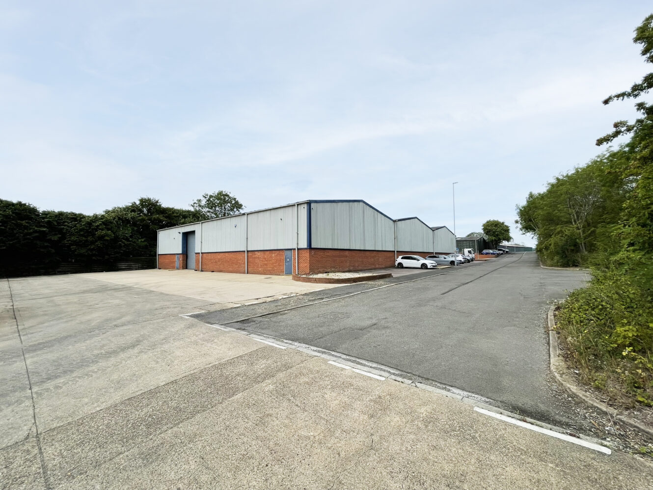 A row of industrial warehouse buildings with brick and metal cladding, adjacent to a tarmacked road and car park with several parked cars. Trees and bushes line the background.