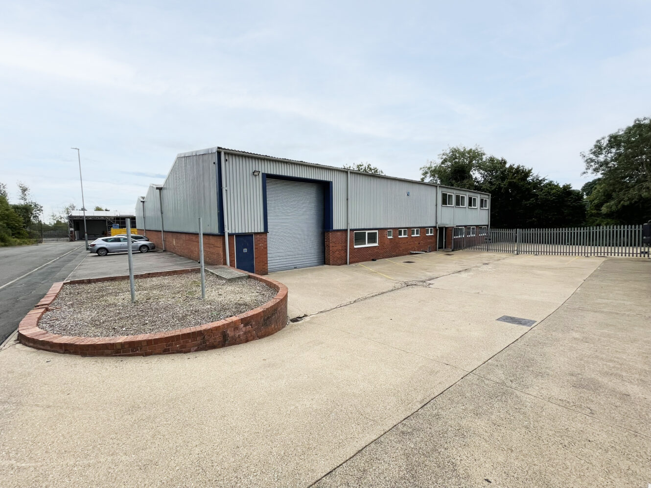 A large industrial warehouse with a grey roller shutter, brick and metal cladding, adjacent parking spaces, and a fenced perimeter under a clear sky.