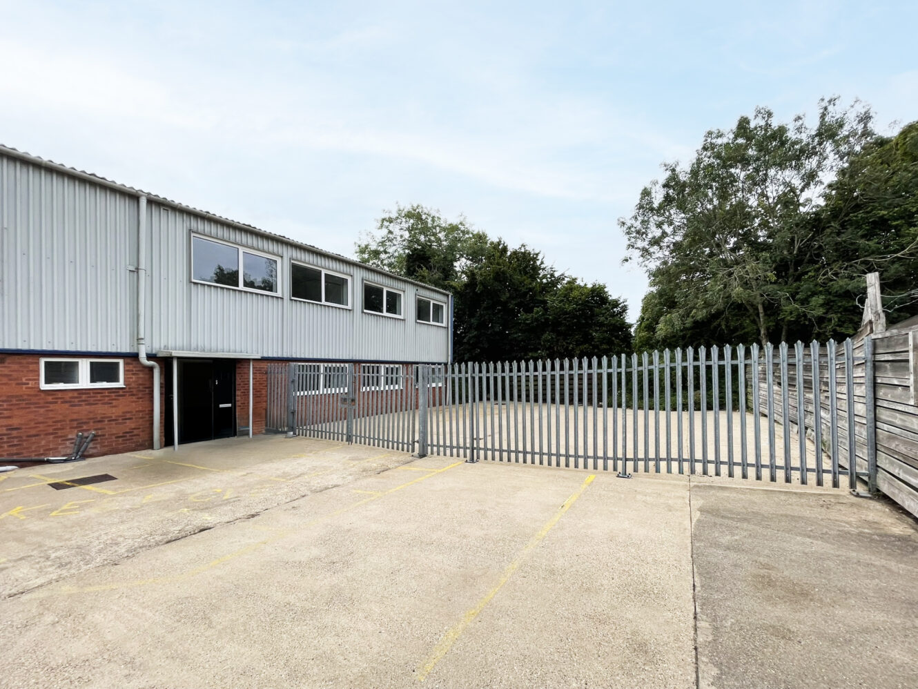 A fenced industrial building with brick and metal cladding, adjacent to an empty tarmacked yard and surrounded by trees.