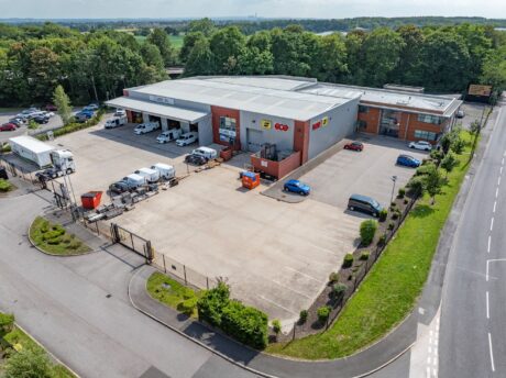 Aerial view of a modern warehouse facility with loading bays, vans, cars, and a fenced car park, surrounded by trees and a main road.