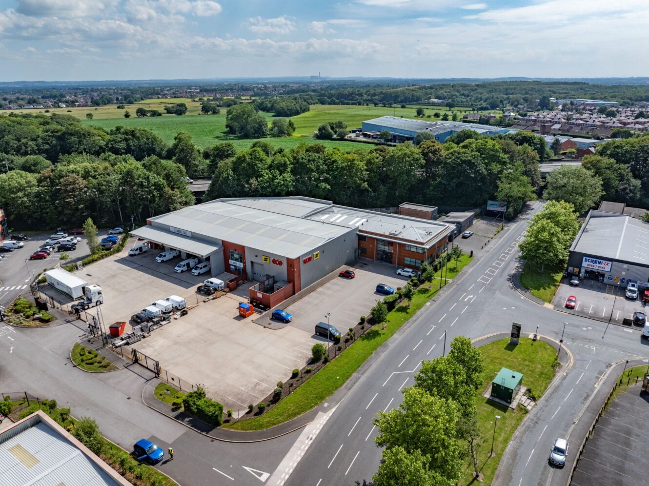 Aerial view of an industrial estate with warehouses, parked vehicles, and surrounding greenery on a clear day.