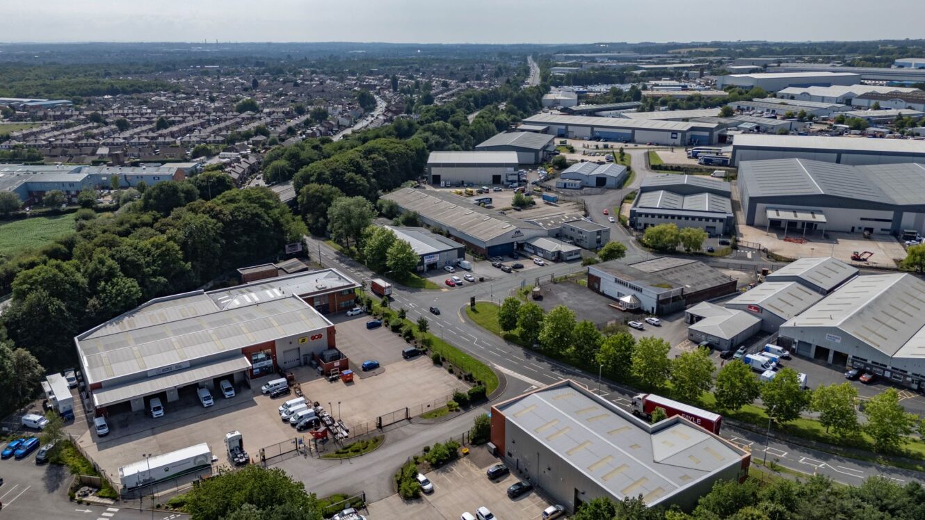 Aerial view of an industrial estate with warehouses, loading bays, parked lorries, and surrounding greenery, with residential areas in the background.