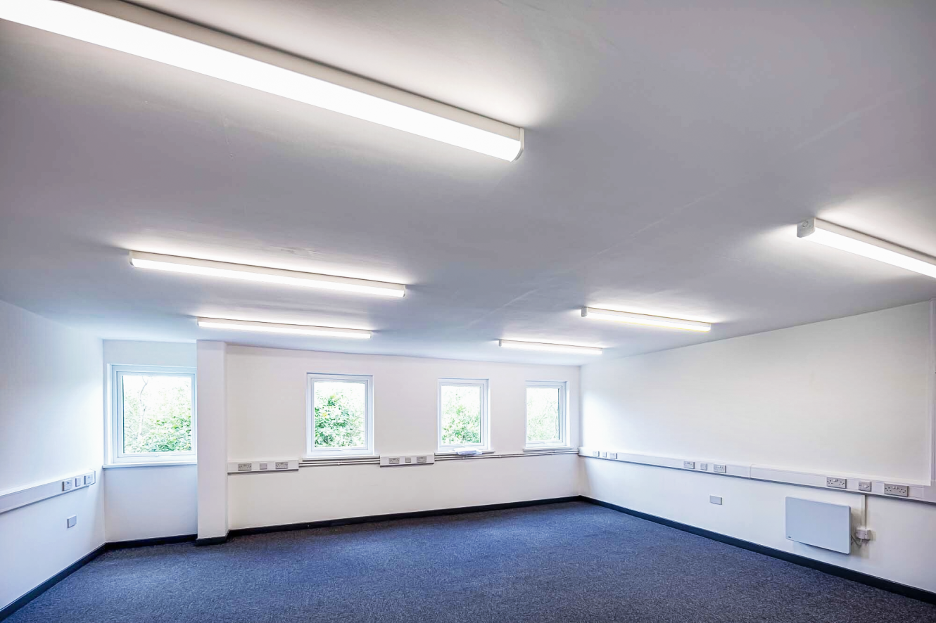 Empty office room with white walls, carpeted floor, several power sockets along the walls, three windows, and fluorescent ceiling lights.
