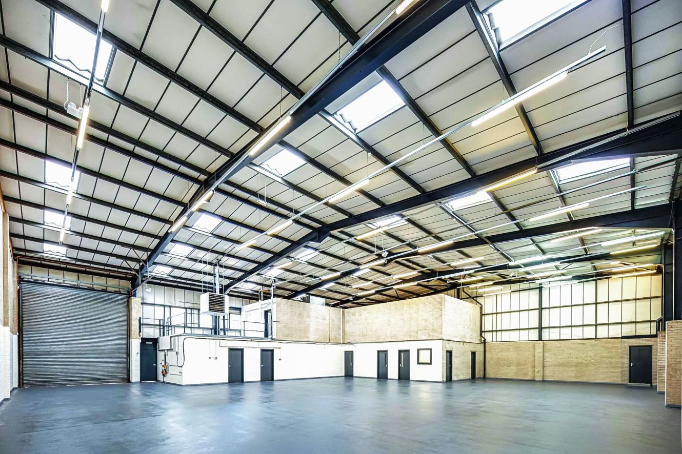 Large, empty industrial warehouse with high ceilings, exposed beams, concrete floor, brick walls, and several doors and windows along one side.