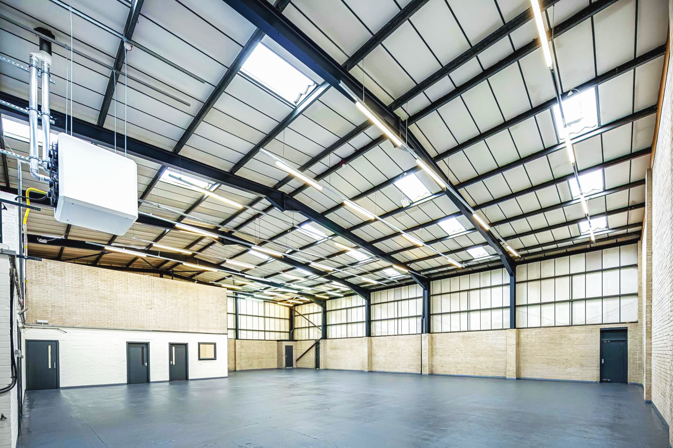 Interior of an empty industrial warehouse with high ceilings, exposed steel beams, skylights, and light-coloured brick walls.