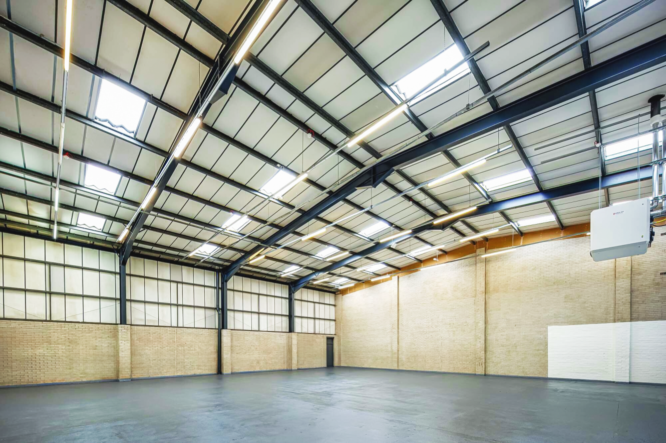 Spacious empty warehouse with high ceilings, metal beams, brick walls, and large overhead rooflights letting in natural light.