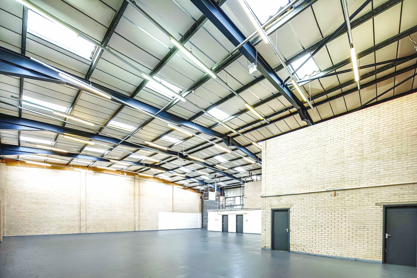 Spacious, empty warehouse interior with high ceilings, exposed steel beams, skylights, brick walls, and several closed doors.