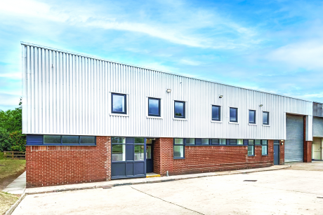 A modern industrial building with a brick lower level, metal upper cladding, multiple windows, and a large roller shutter, set under a blue sky.