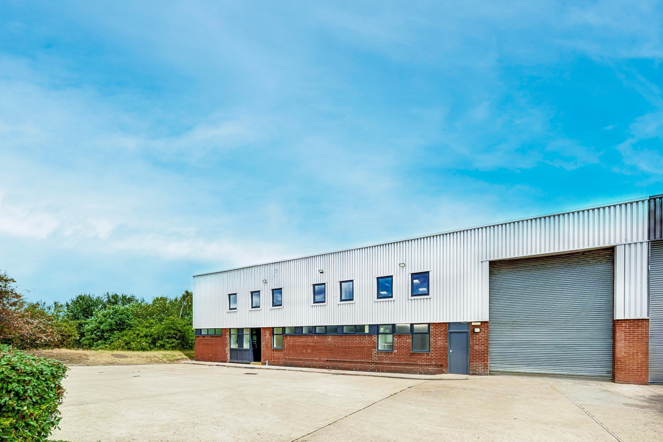 A large industrial warehouse with a brick and metal exterior, several small windows, and a closed roller shutter, set against a blue sky.