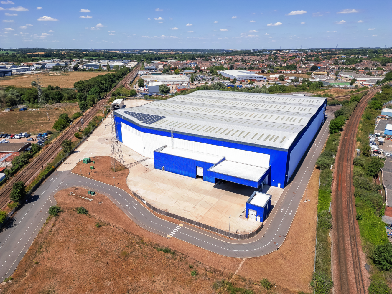 A large blue and white industrial warehouse with loading bays and paved areas, surrounded by roads and railways, sits in a semi-rural area under a sunny sky.