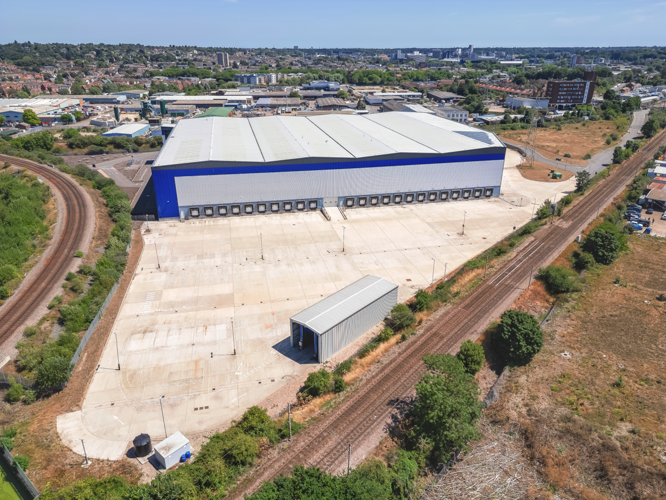 A large industrial warehouse with a blue and white exterior sits beside a railway track and a spacious concrete loading area, surrounded by greenery and urban buildings in the background.