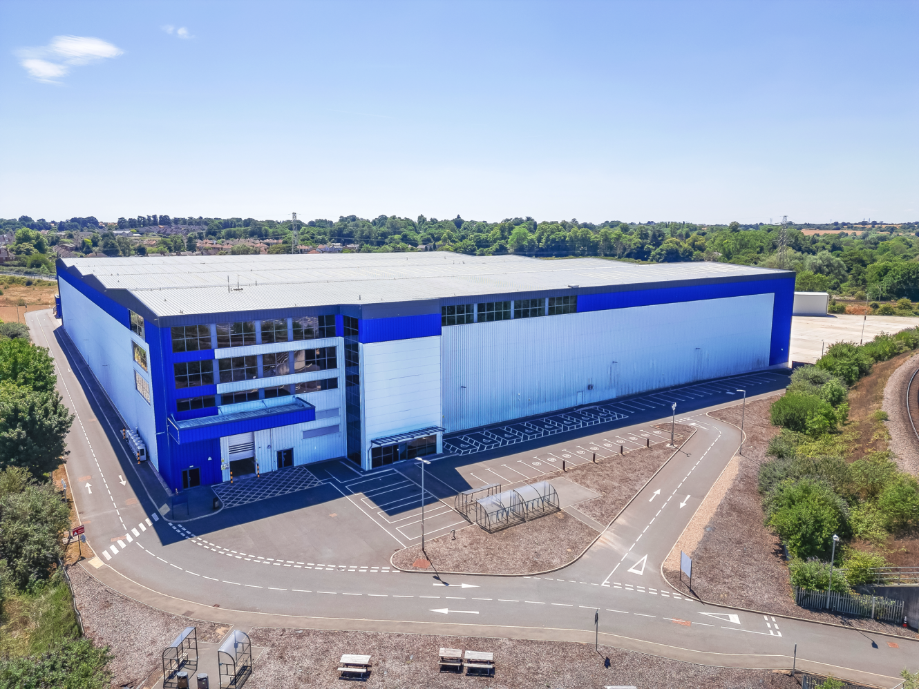 A large, modern warehouse or industrial building with blue and white exterior, surrounded by empty parking spaces and landscaping under a clear sky.