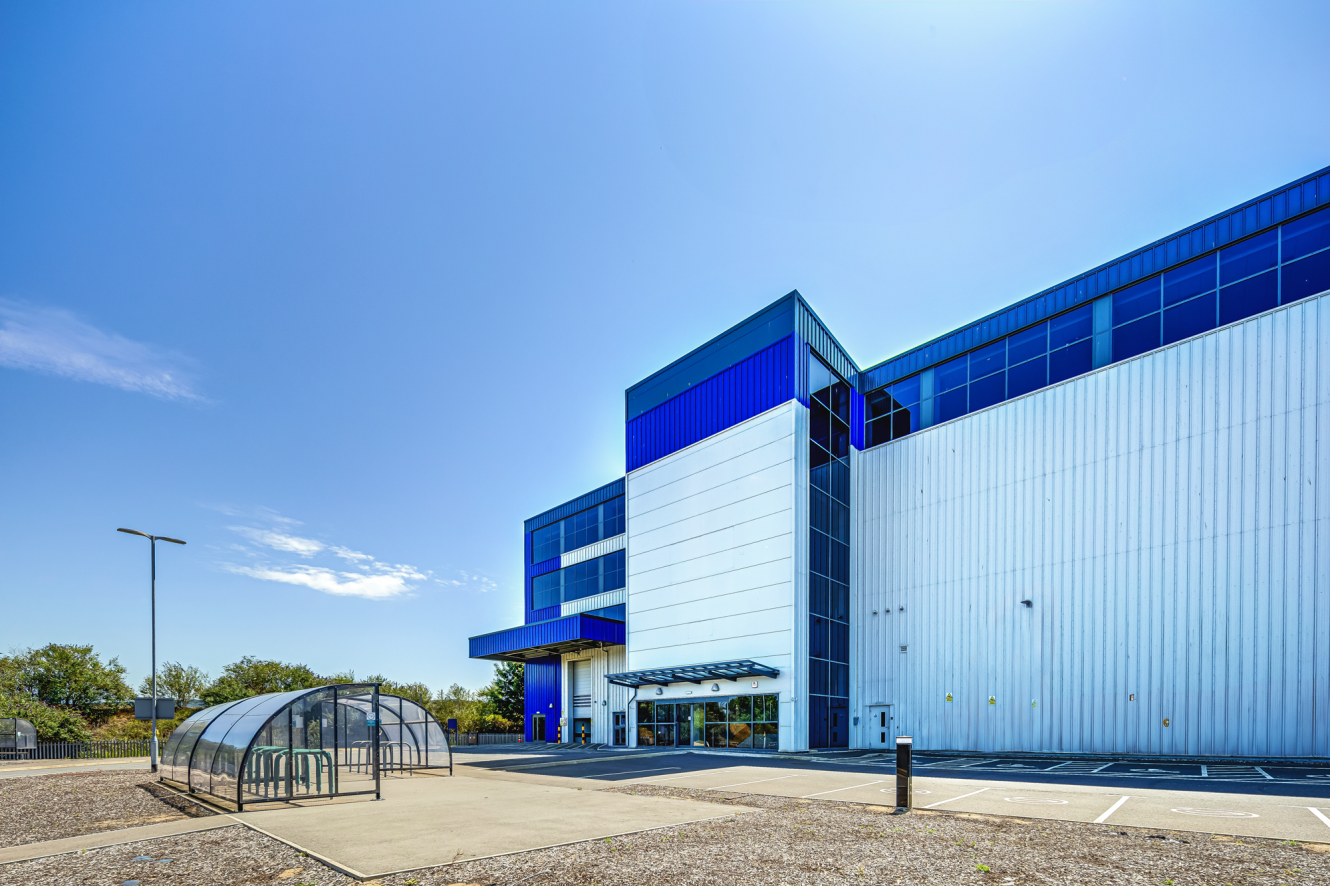 Modern industrial building with blue accents, large windows, and a covered bike shelter, set against a clear blue sky with an empty car park in front.