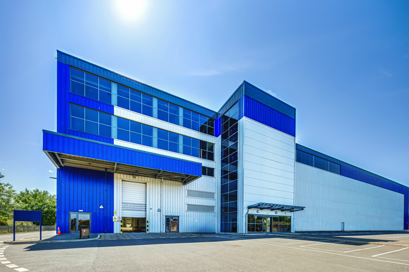 Modern industrial warehouse building with blue and white panels, large glass windows, and loading bay doors under a clear sunny sky.