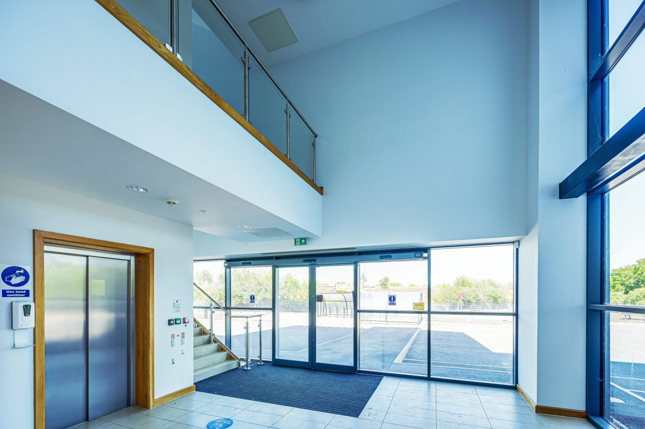 Modern building entrance with glass doors, a lift, a staircase with a glass balustrade, and large windows allowing natural light into the lobby area.