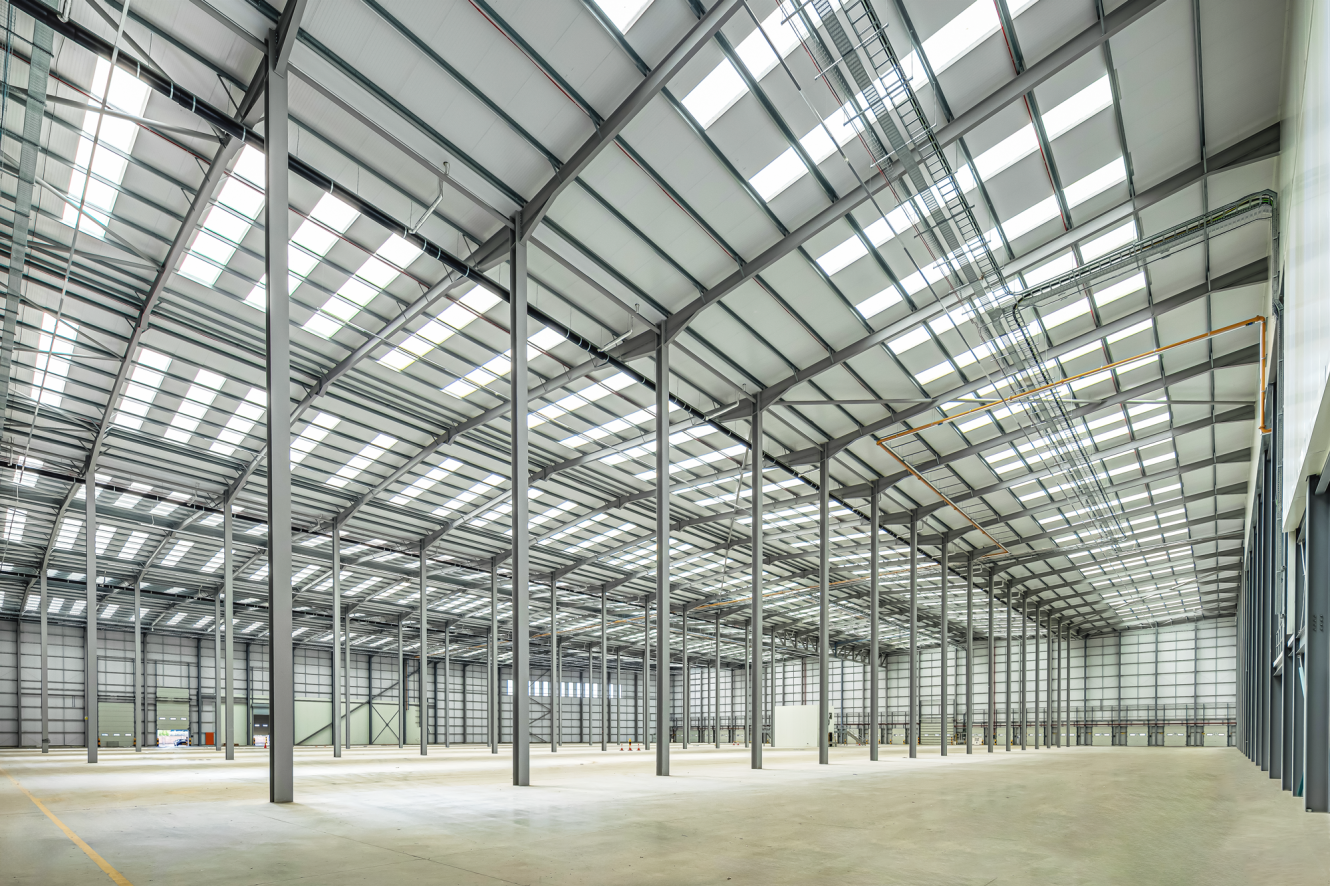 Interior view of a large, empty warehouse with high ceilings, metal beams, and skylights allowing natural light in.