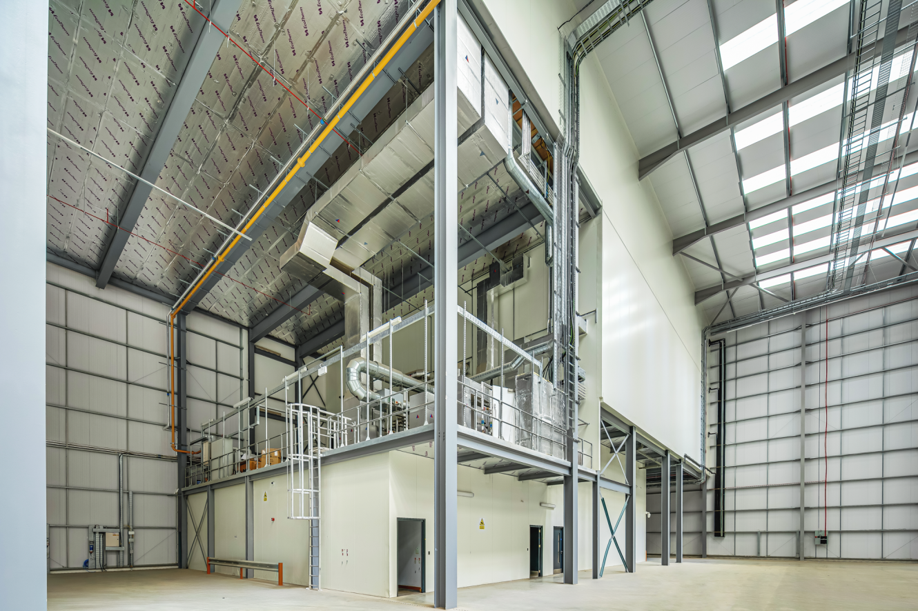 Interior of a large, modern industrial facility with metal beams, insulated ceiling, ventilation ducts, and an elevated platform structure above a white-walled room.