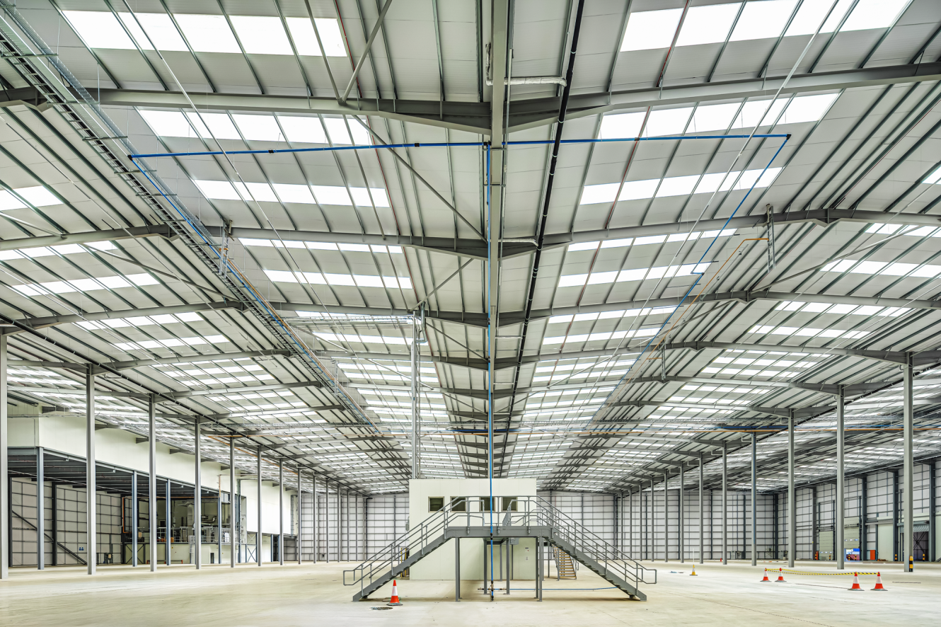 Interior of a large, empty warehouse with a small white office structure and staircase in the centre, surrounded by metal beams and skylights.