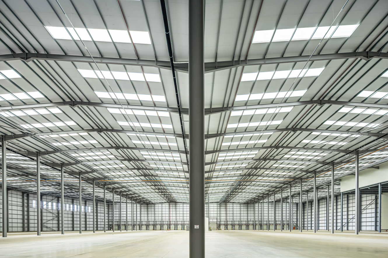 Interior of a large, empty warehouse with high ceilings, exposed beams, and evenly spaced support columns under bright overhead rooflights.