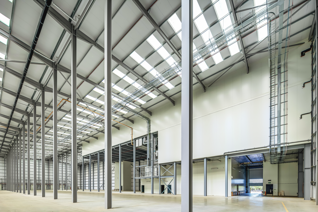 Large, empty industrial warehouse with high ceilings, exposed beams, skylights, concrete floor, and open roller doors allowing natural light inside.